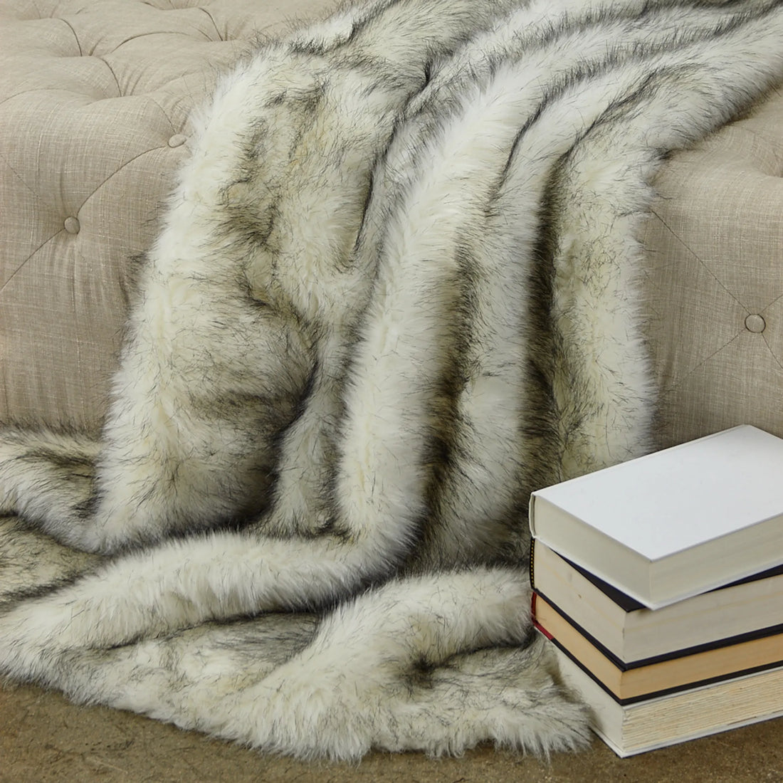 Gray fur blanket draped over a beige sofa with a stack of books on a wooden floor.