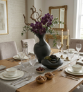 Elegant dining table setting with purple flowers in a dark vase, candles, and small dishes.