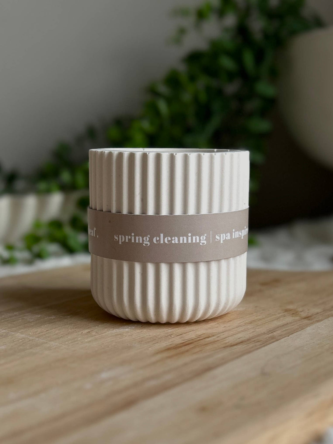 White ribbed candle with a gray label on a wooden surface with greenery in the background