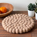 Round woven mat on a wooden surface with a bowl of oranges and potted plants in the background.