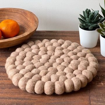 Round woven mat on a wooden surface with a bowl of oranges and potted plants in the background.