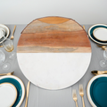 Round wooden and white placemat on a gray surface with dinnerware and cutlery.